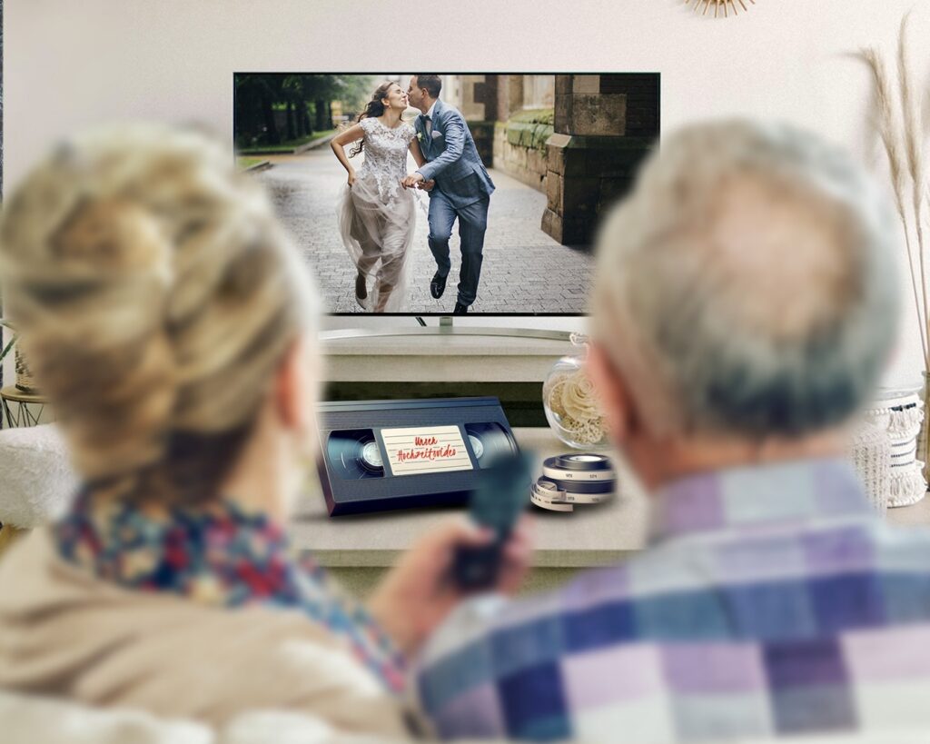 ein glückliches paar sitz im Wohnzimmer vor TV Bildschirm und sieht sich ihren Hochzeitfilm an. Davor liegt die originale VHS Videokassette.